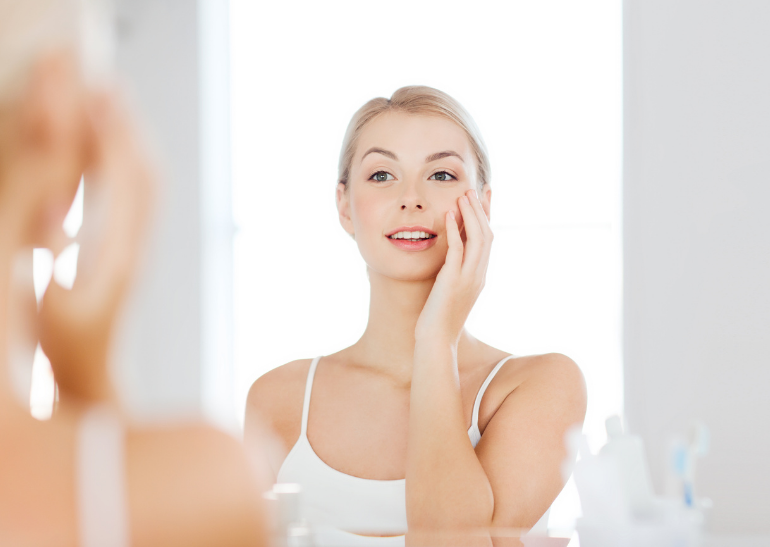 woman looking in the mirror, one hand on face admiring her healthy skin