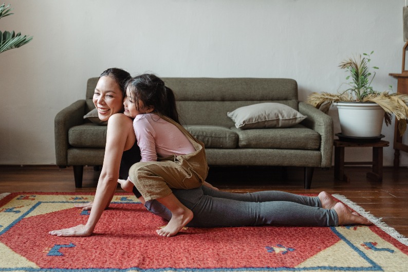 mom practicing yoga, smiling over her shoulder at daughter who is sitting on her back, hugging her from behind