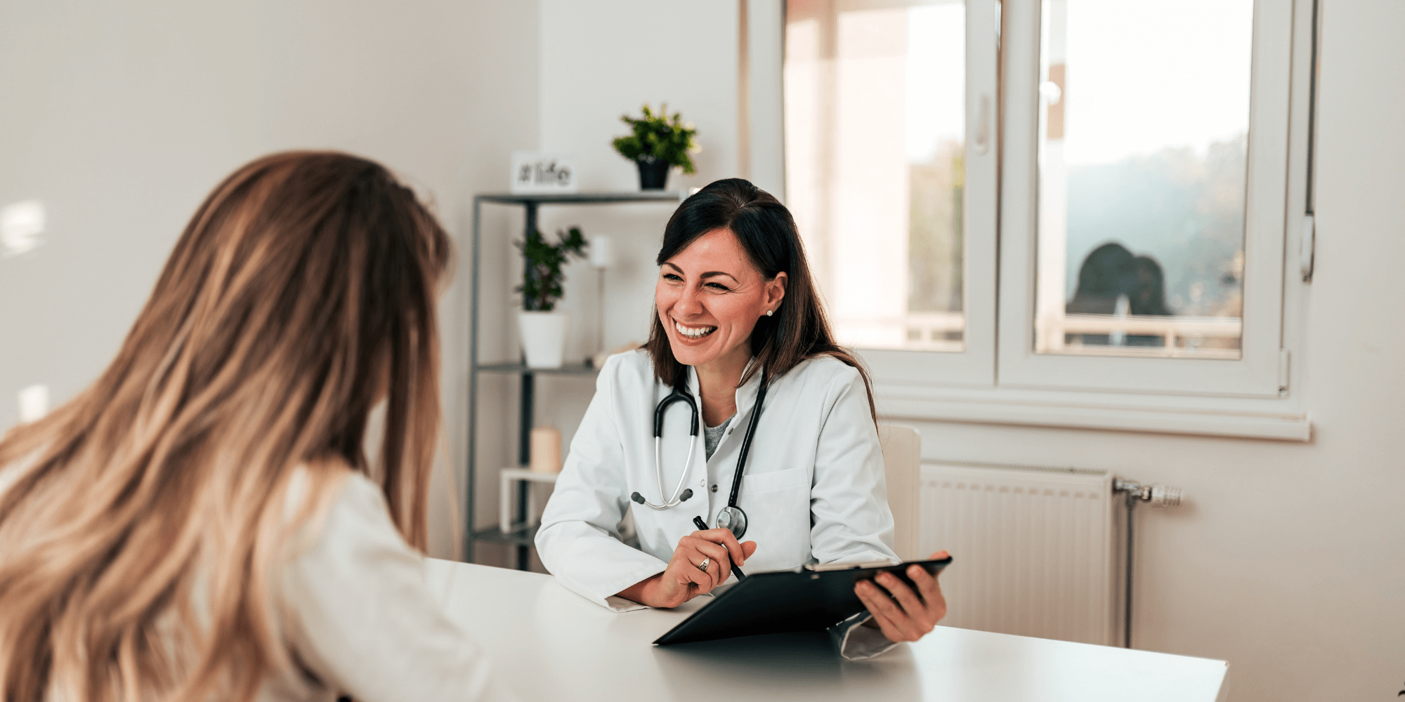 Female doctor sitting at a table in an office,  holding a clipboard smiling at a female patient